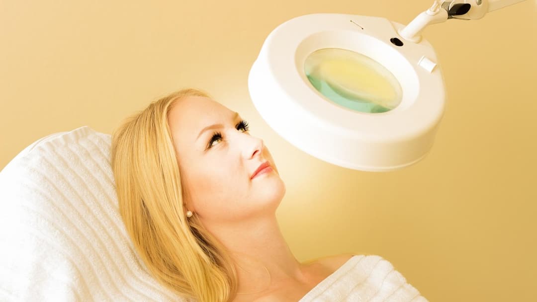 Blonde woman with doll lashes and sculpted facial features under a cosmetic clinic magnifying lamp