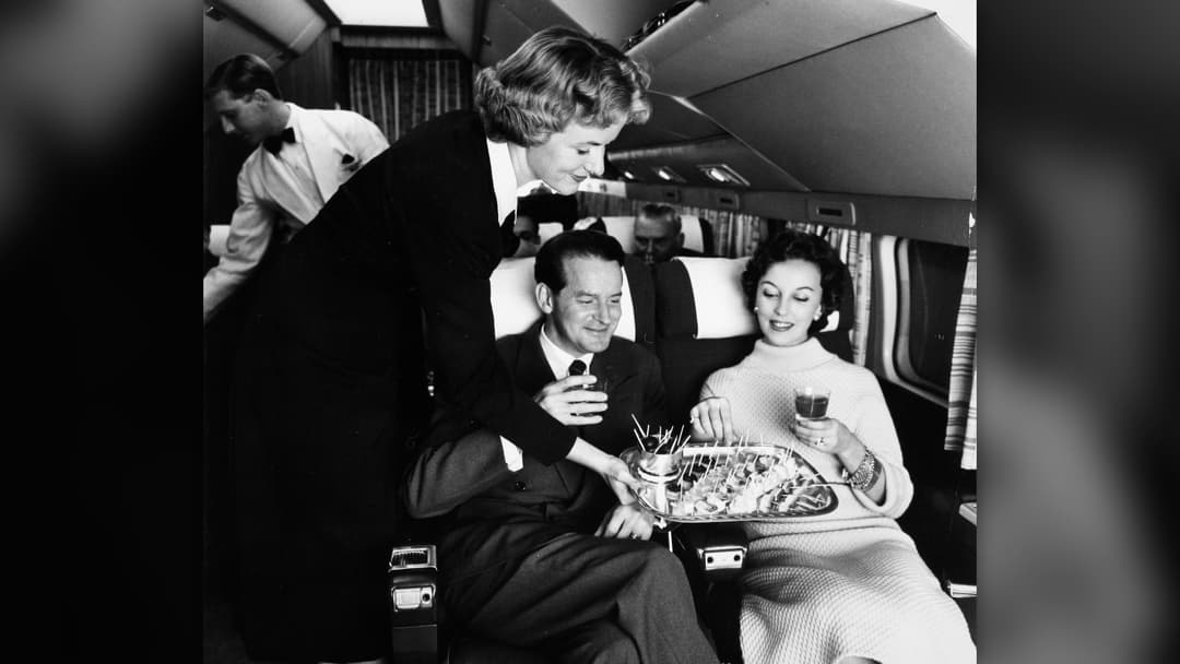 Vintage mid-century air hostess serving food and snacks to seated passengers onboard an airliner