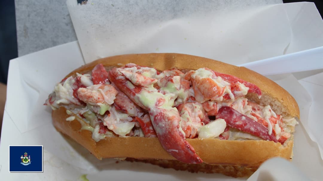Overstuffed traditional Maine lobster roll on a toasted split-top bun with Maine flag overlay in bottom-left corner