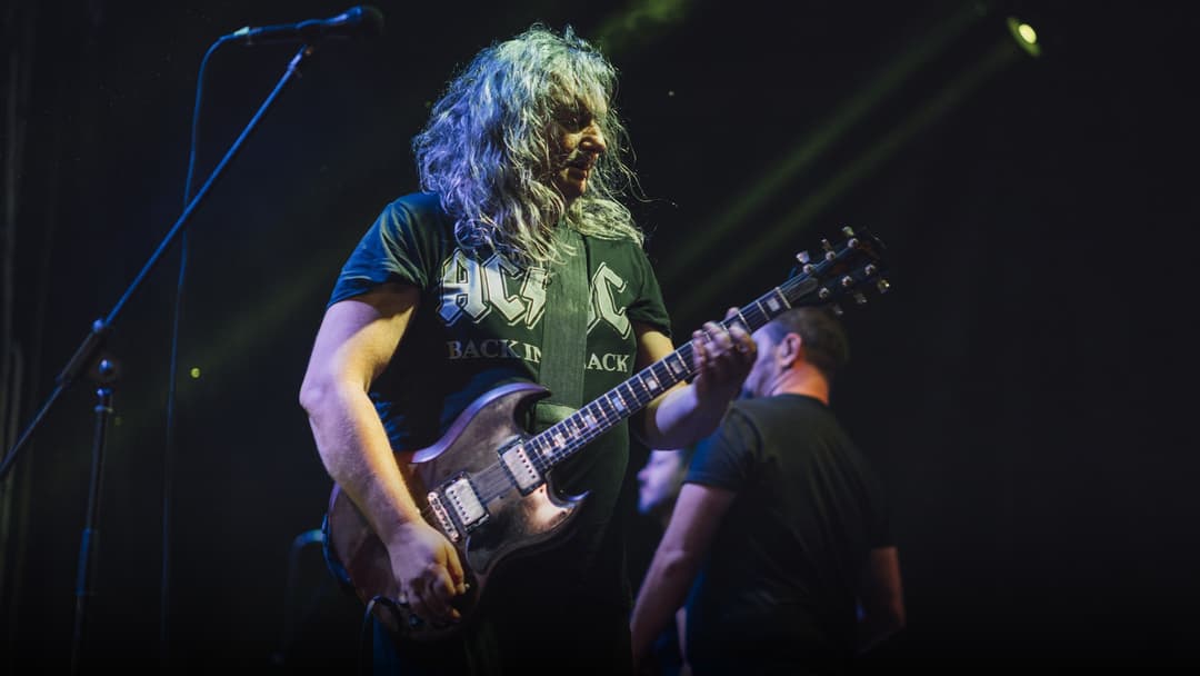 A rock musician performing on an electric guitar under stage concert lighting.