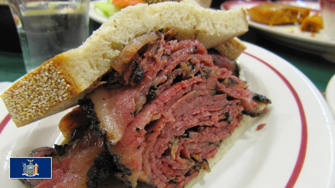 Massive classic New York deli pastrami sandwich on rye bread with New York flag overlay in bottom-left corner