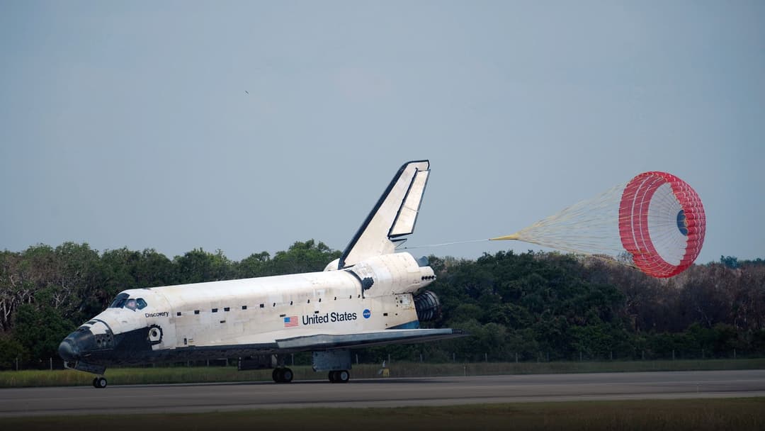 Space Shuttle Discovery touching down on a military airfield runway with red and white drag chute deployed.