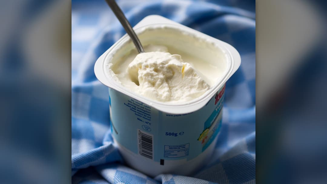 An opened tub of thick white strained yogurt with a metal spoon resting inside.