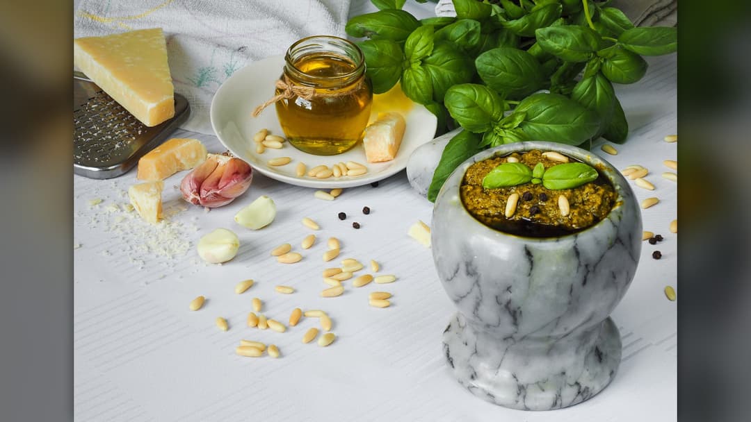 A white marble mortar filled with green basil pesto beside pine nuts, garlic cloves, Parmigiano cheese, and a jar of olive oil.