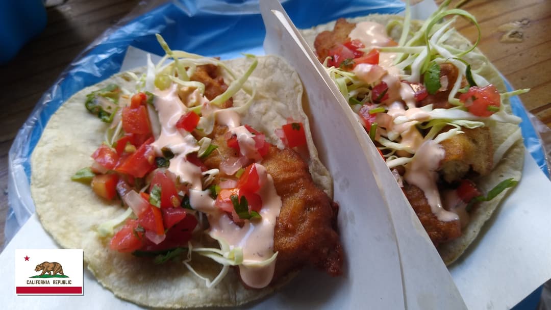 Platter of fresh Baja-style crispy fish tacos with lime wedges and California flag overlay in bottom-left corner