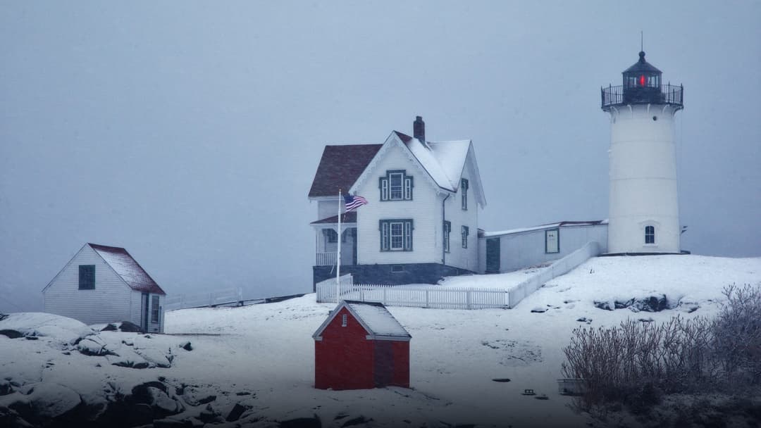 Cape Neddick Lighthouse standing atop a snowy coastal rock bluff overlooking the ocean.