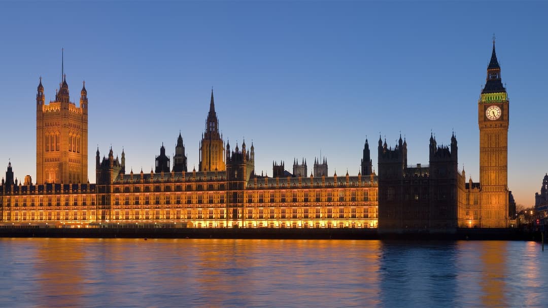 The illuminated Gothic facade of the Palace of Westminster and Big Ben clock tower reflected across the River Thames at dusk.