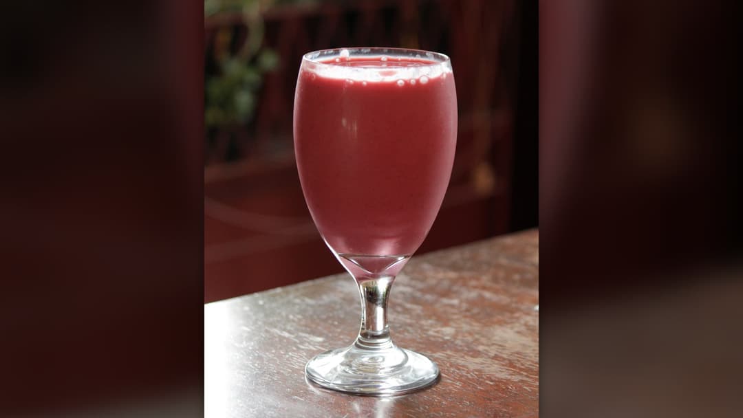 A stemmed glass filled to the brim with thick pink strawberry fruit smoothie on a wooden table.