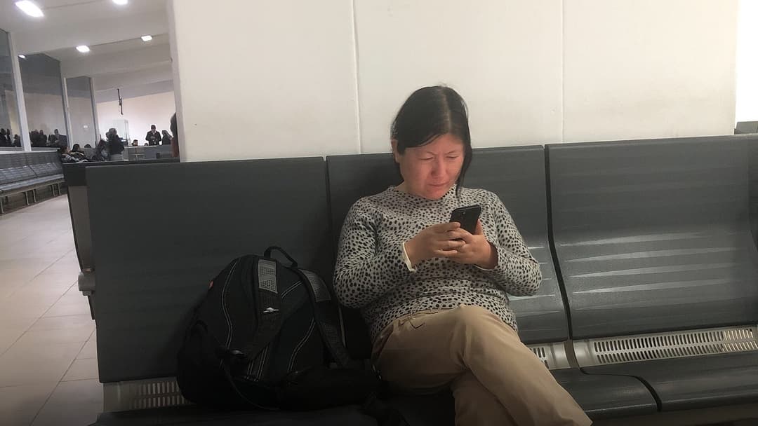 Passenger seated on airport terminal gate chairs looking intently at a smartphone screen without headphones.