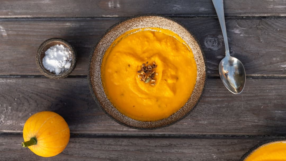 Overhead view of a warm bowl of orange pumpkin soup garnished with seeds on a wooden table.