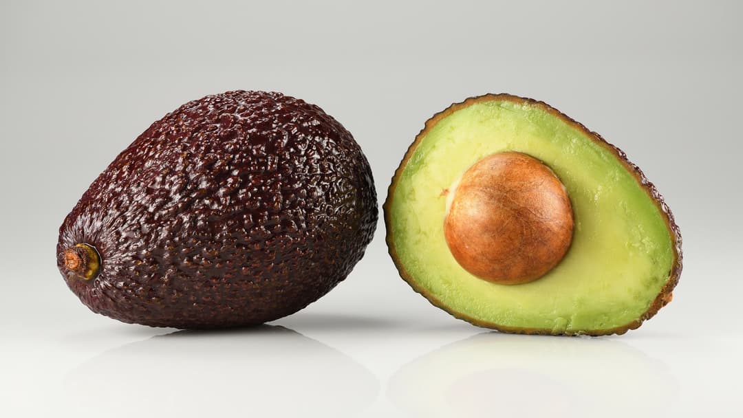 A whole dark-skinned Hass avocado next to a halved Hass avocado showing its green flesh and central pit.