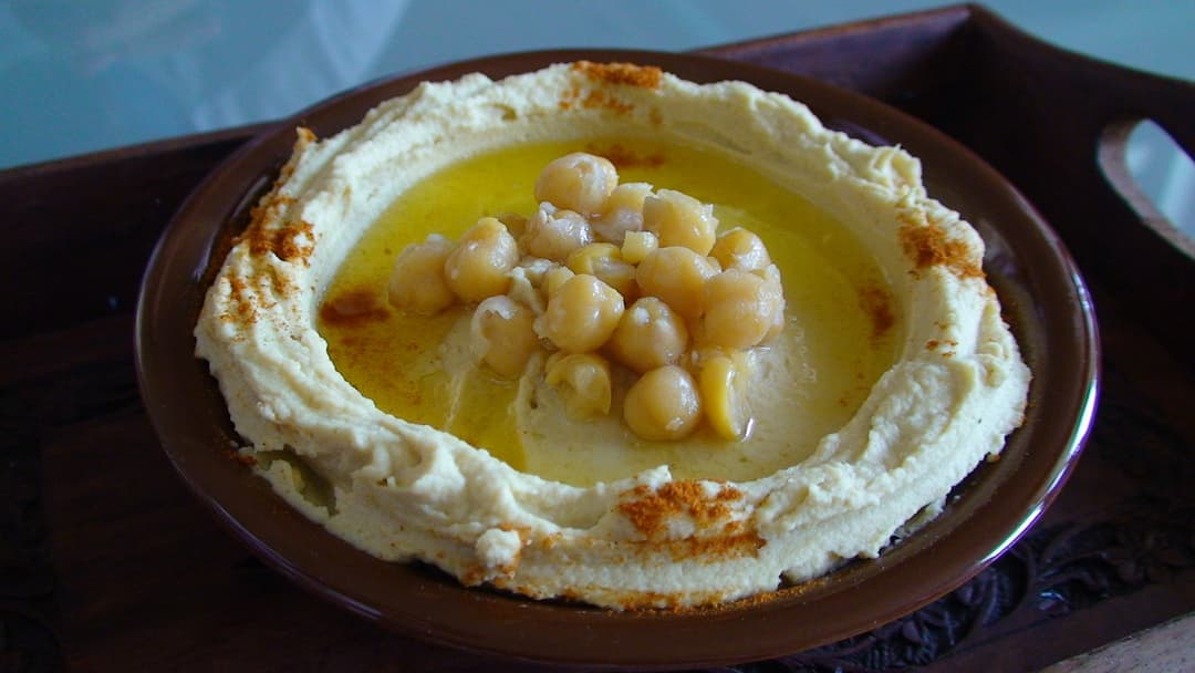 A brown ceramic bowl of creamy chickpea hummus topped with whole chickpeas and a pool of golden olive oil.