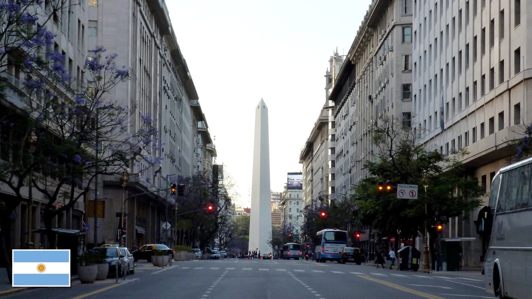 The Obelisco framed by blooming purple jacarandas in Buenos Aires, Argentina with the Argentine flag in the bottom left corner