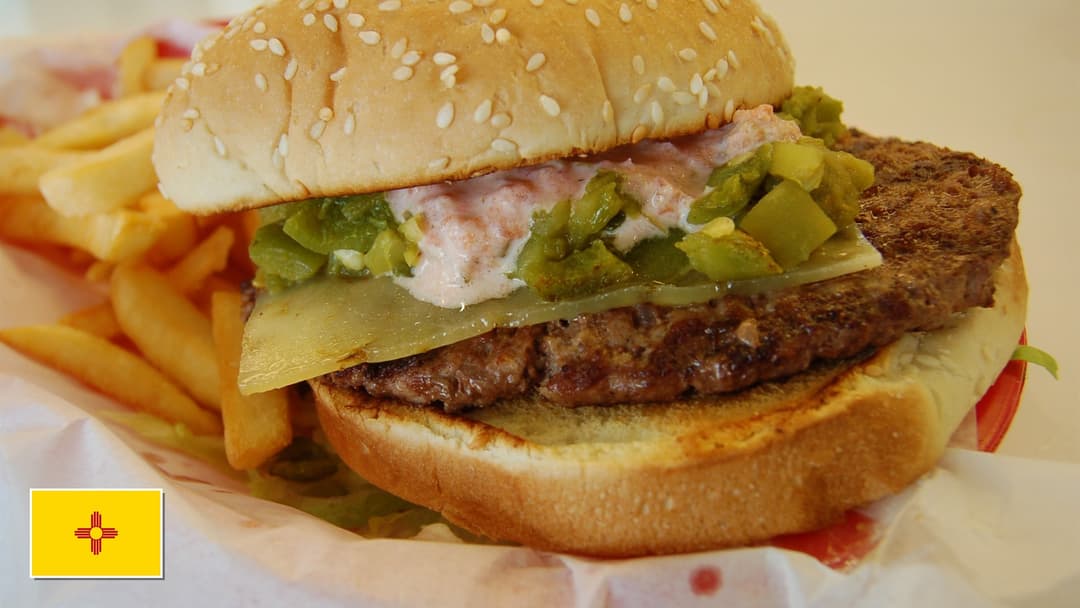 Juicy burger topped with melted cheese and fire-roasted Hatch green chiles with New Mexico flag overlay in bottom-left corner