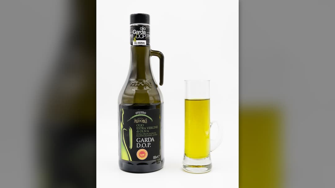 A dark glass bottle of Garda DOP extra-virgin olive oil standing next to a clear glass mug filled with golden olive oil.