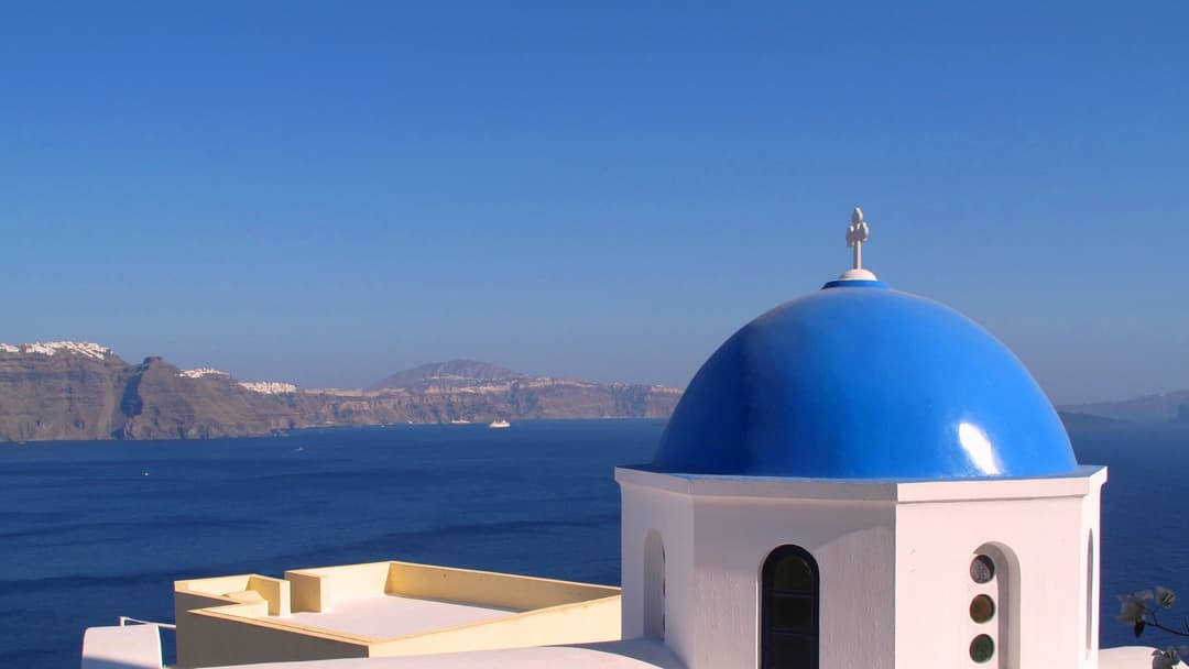 Iconic blue-domed white churches of Oia overlooking the sunlit Aegean caldera in Santorini.