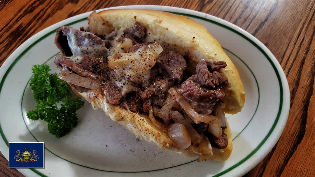 Freshly grilled authentic Philly cheesesteak overflowing with shaved beef and melted cheese with Pennsylvania flag overlay in bottom-left corner