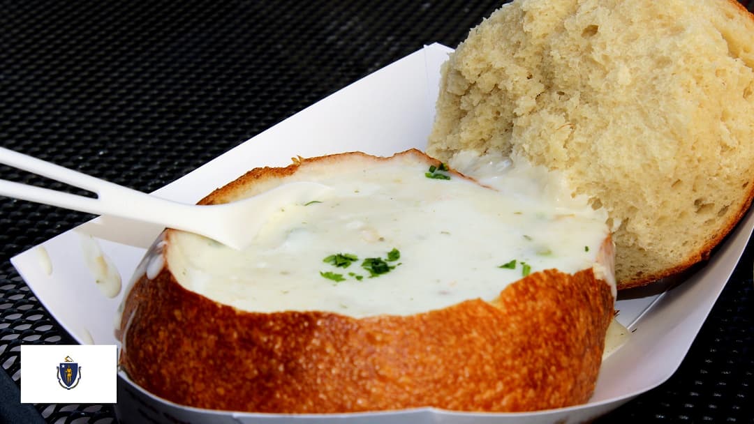 Steaming bowl of creamy New England clam chowder in a sourdough bread bowl with Massachusetts flag overlay in bottom-left corner