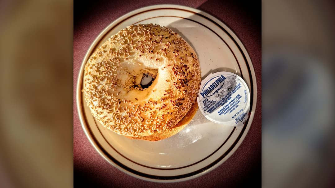 A toasted sesame seed bagel served on a ceramic diner plate beside an individual cup of cream cheese.