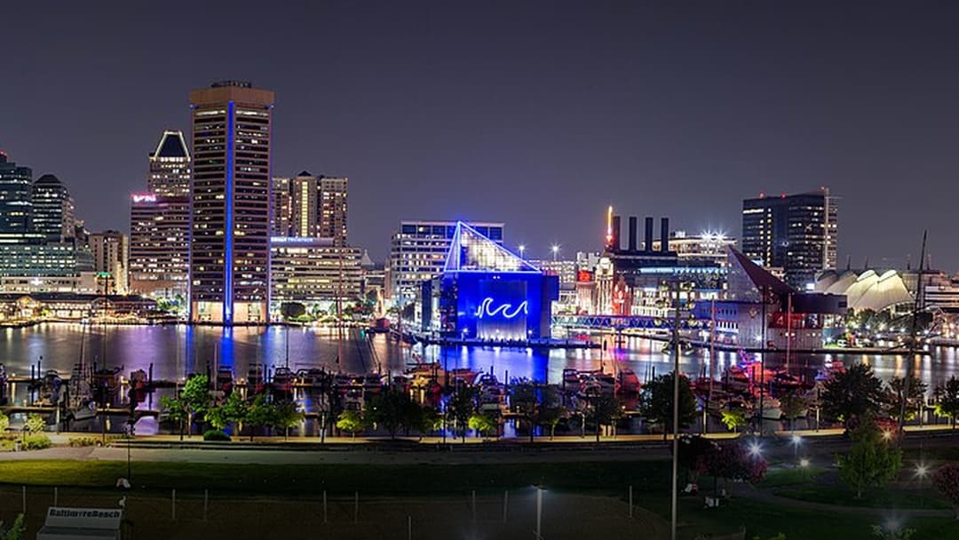 Illuminated nighttime cityscape of Baltimore's Inner Harbor with water reflections.