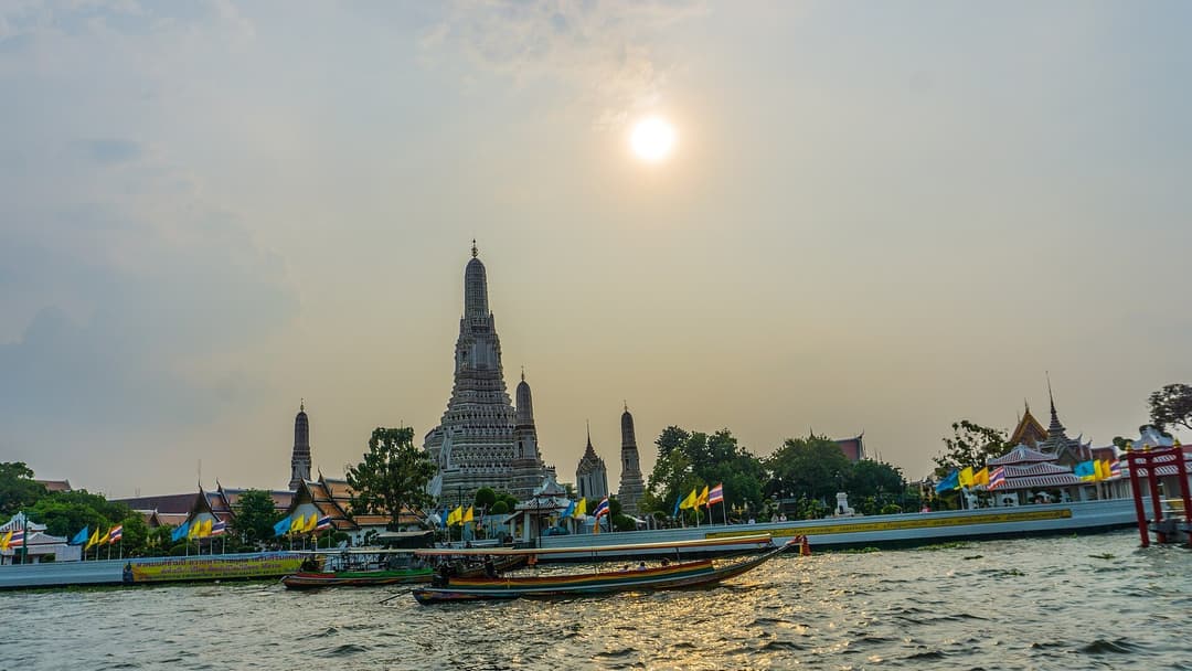 Wat Arun Temple of Dawn with its iconic central prang tower and longtail boats on the Chao Phraya River at sunset.