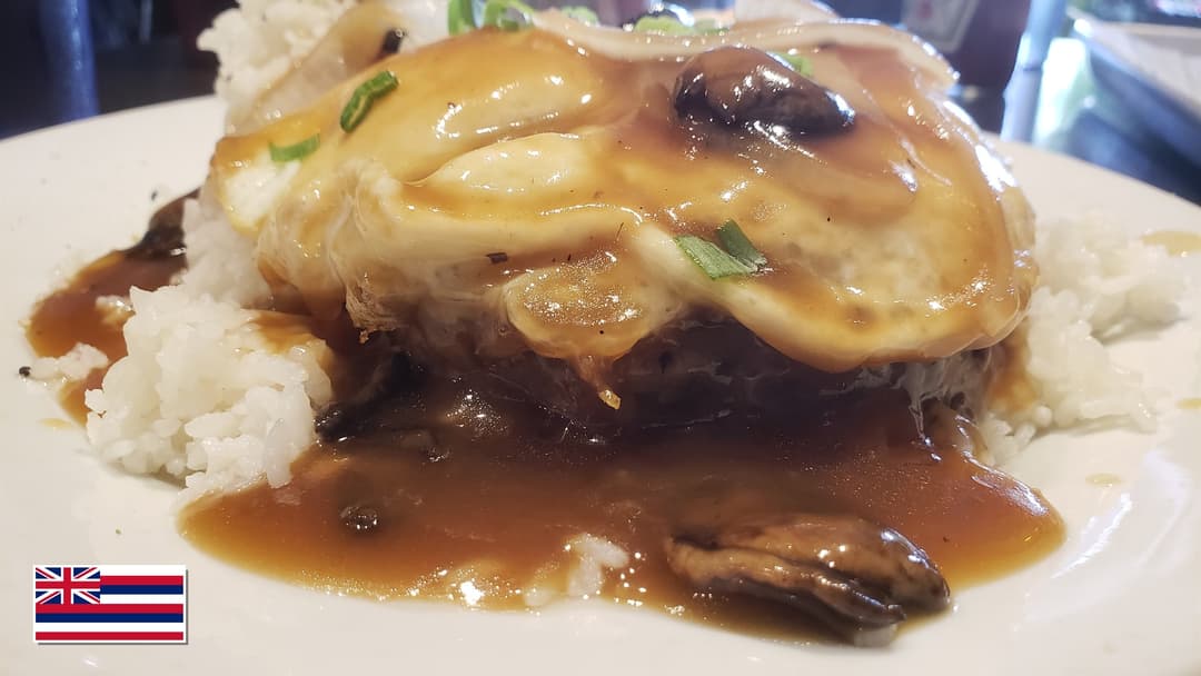 Hawaiian loco moco with grilled beef patty, sunny egg, and brown gravy over rice with Hawaii flag overlay in bottom-left corner