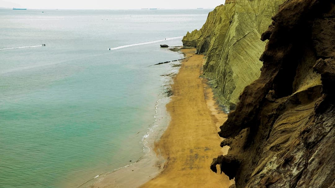 Coastal cliffs and ochre shoreline of Hormuz Island overlooking ships in the Strait of Hormuz.