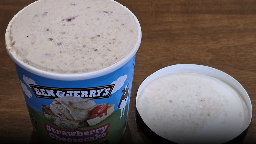 An open pint of Ben and Jerry's Strawberry Cheesecake ice cream with the colorful illustrated lid resting behind it.